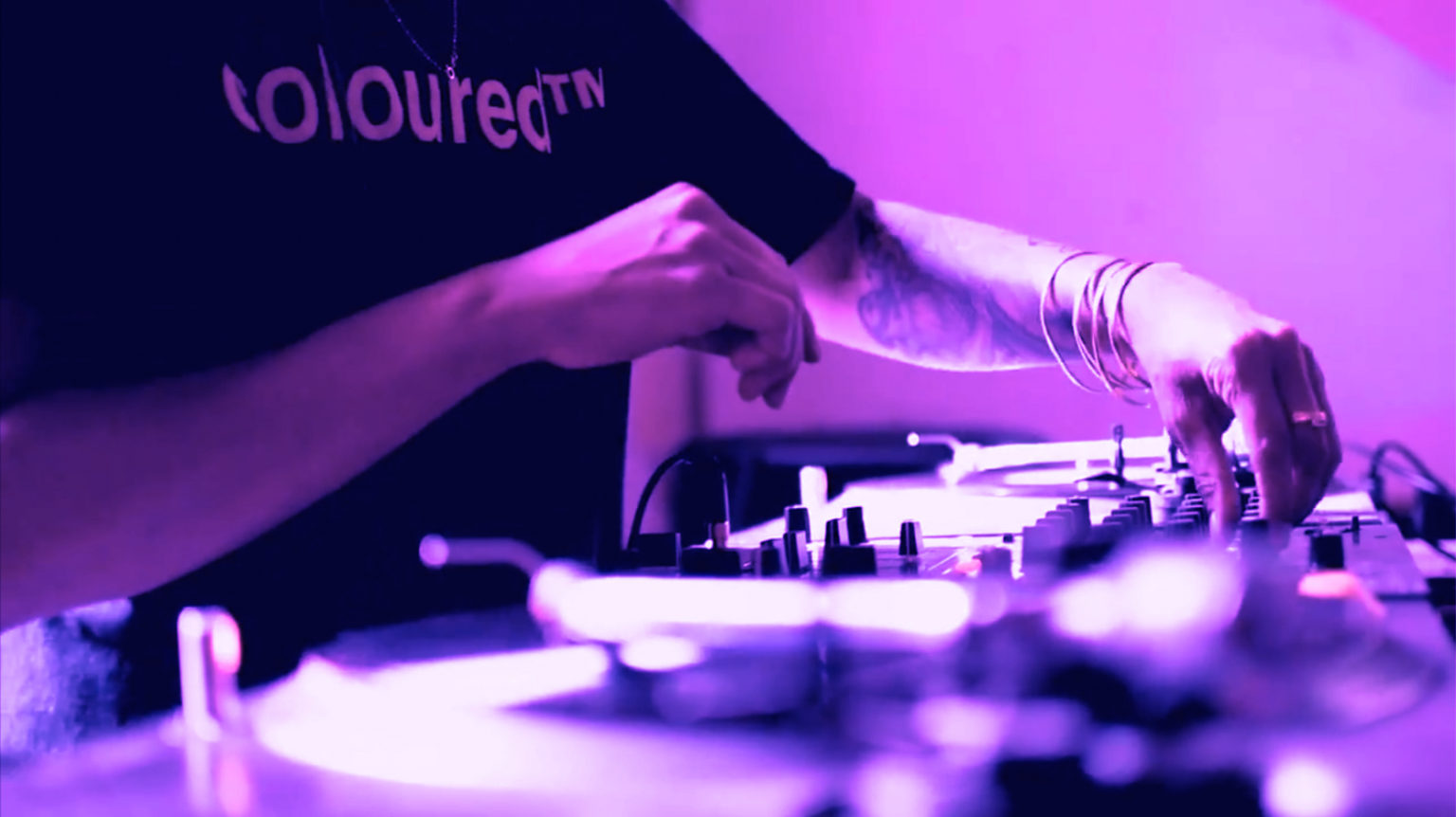 An image of a DJ turning knobs on a mixer, wearing multiple bangles and Roberta Joy Rich’s Coloured™ T-Shirt.