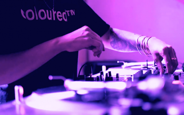 An image of a DJ turning knobs on a mixer, wearing multiple bangles and Roberta Joy Rich’s Coloured™ T-Shirt.