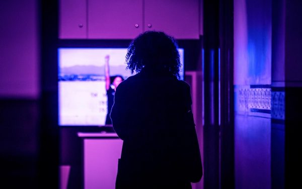 An image of an audience member wearing a large, dark trench coat with short curly hair stands in a dimly lit hallway, viewing a screen that is out of focus with the image of a person holding the Black Power fist salute in a brightly lit outdoor space.