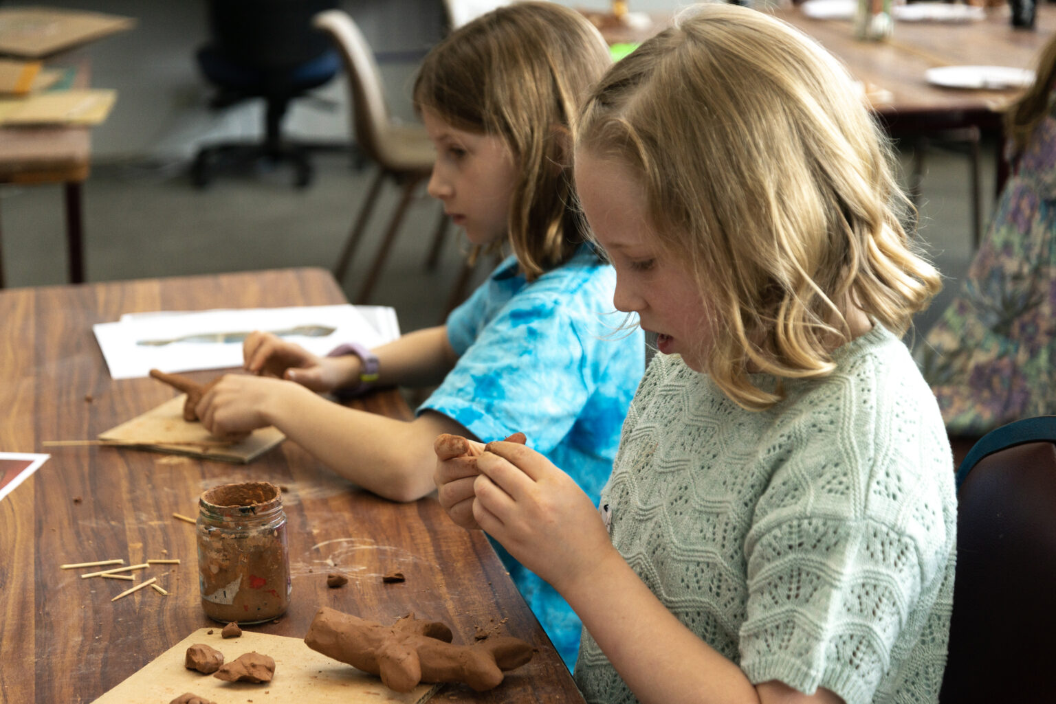 We see two kids sitting at a wooden table concentrating on creating clay animals.