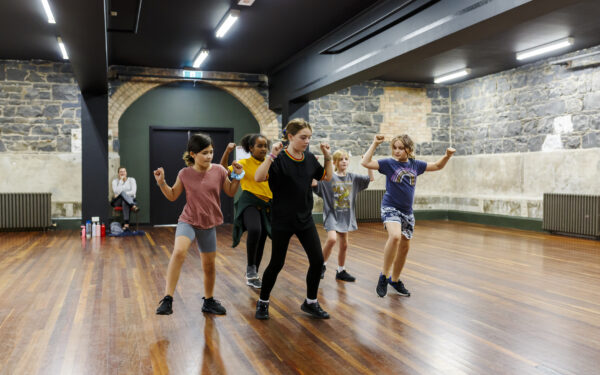 We see a room with wooden floors and bluestone walls. There are a group of kids in the middle all dancing with their hands in the air.