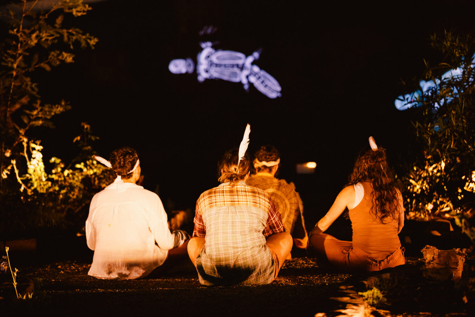 Four people are sitting outdoors on the ground, wearing headbands with a single feather sticking out. They are looking up at a projection art of a geometric platypus in the night sky above them. The image has earth tones and is taken at nighttime.