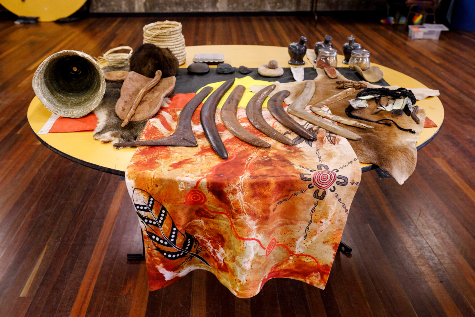 A table is shown with a variety of First Nations tools and instruments placed on top of a patterned material and possum skin. Among these items are several boomerangs, which are wooden tools that are curved in a unique shape and used for hunting or sport. Other tools include smooth rocks and woven baskets. The patterned material underneath the objects features bold geometric shapes and vibrant colours, while the possum skin adds a layer of texture and depth to the overall display.